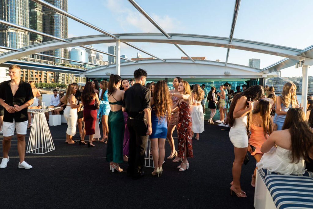 A group of people socializing at a rooftop event with city buildings in the background. The attendees, dressed in semi-formal and summer outfits, are mingling around high tables and enjoying the sunny weather. The ambiance resembles that of The Yacht Social Club's boat parties on Sydney Harbour.