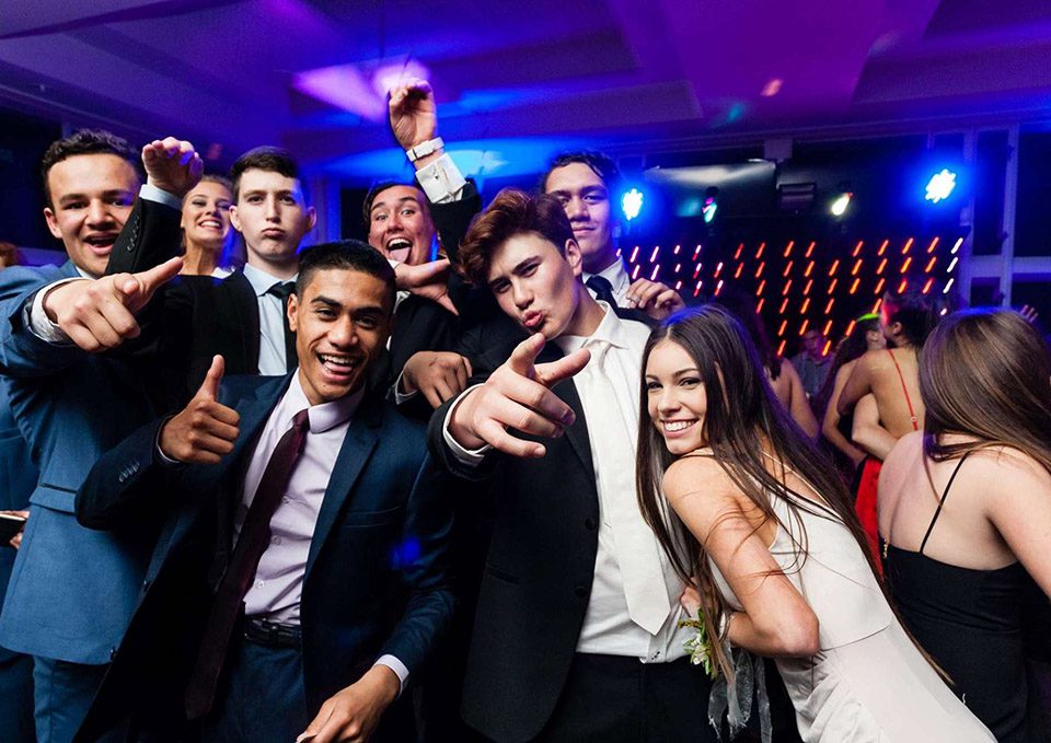 A group of young people dressed in formal attire are posing energetically at a party, pointing towards the camera and smiling widely. The background features colorful lights and a lively atmosphere, reminiscent of an exclusive gathering by Sydney Harbour Boat Hire The Yacht Social Club.