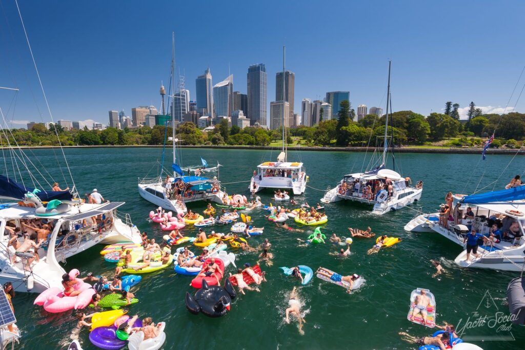 A lively scene of boats gathered in a harbor with people enjoying the water on colorful inflatables and swimming. The Sydney CBD skyline with tall buildings and lush green trees provides a vibrant backdrop under a clear blue sky.