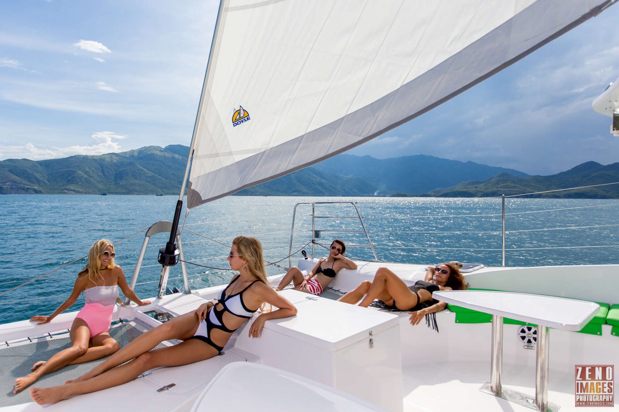 Four people relax on a sailboat under a clear sky. Three women in swimsuits and sunglasses are sitting and reclining on the boat's deck, while a man in swim trunks lounges near the edge. The background features calm blue water and distant mountains.