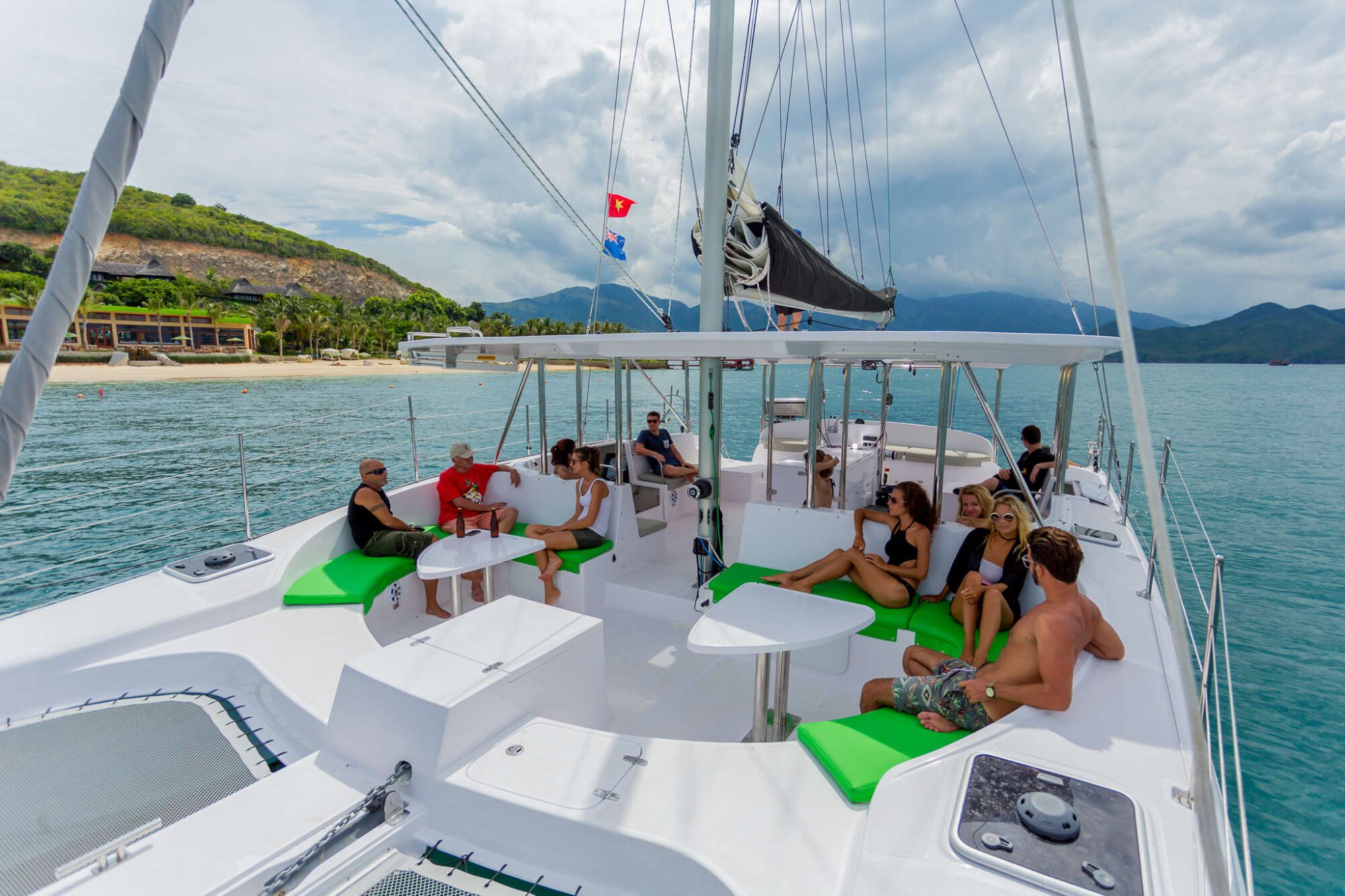 A group of people relaxes on a white sailing catamaran with bright green cushions. The boat is sailing in a tropical location with clear blue water and hills in the background. The sky is cloudy, and a shoreline with a few structures is visible nearby.