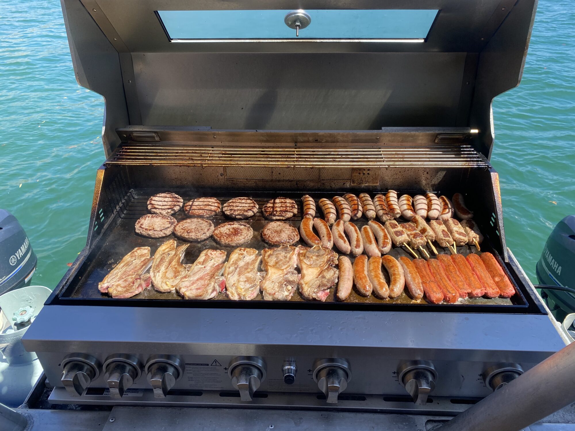 A grill loaded with a variety of meats, including steaks, sausages, burger patties, hot dogs, and skewered items. The grill is positioned by the water, creating a scenic outdoor cooking setting.