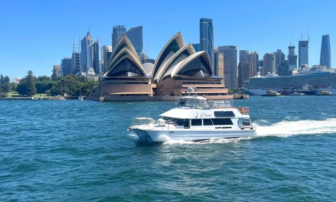 A white boat cruises on a bright blue harbor with the iconic Sydney Opera House and modern city skyline in the background. Clear, sunny skies enhance the vibrant scene, perfect for Luxury Yacht Rentals Sydney or Boat Parties by The Yacht Social Club.