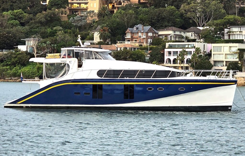 A sleek, blue and white motor yacht floats on water with a backdrop of upscale houses and greenery on a hillside. The yacht has a modern design with large windows and a spacious deck area.