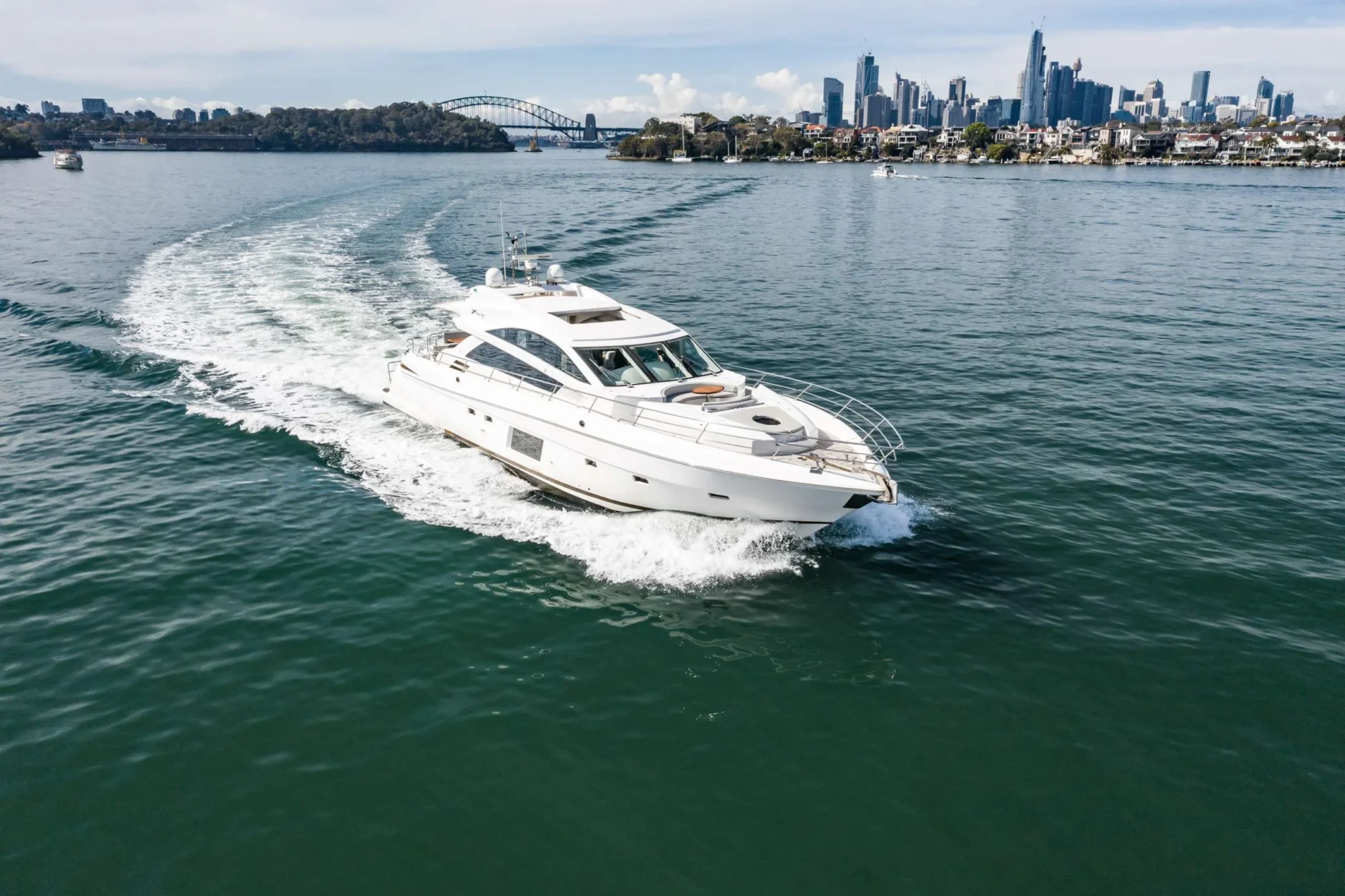 A white yacht cruises on blue water, leaving a wake behind it. In the background, a city skyline is visible with tall buildings and a bridge. The day is clear with minimal clouds. Experience this scene with The Yacht Social Club Sydney Boat Hire for an unforgettable adventure.