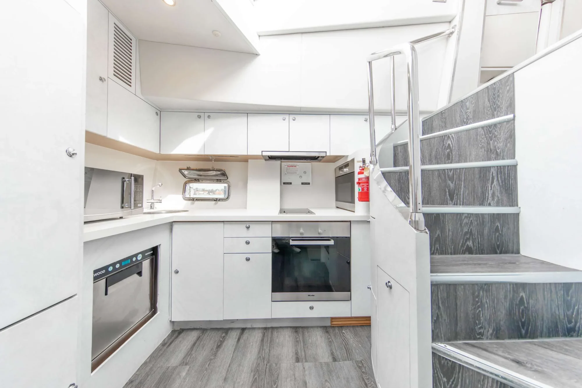 A modern kitchen with white cabinets, an oven, a stovetop, a microwave, and a dishwasher. There's a small window and grey wood flooring. A staircase with metal railings ascends to the right of the kitchen. Perfect for hosting Boat Parties Sydney The Yacht Social Club events.