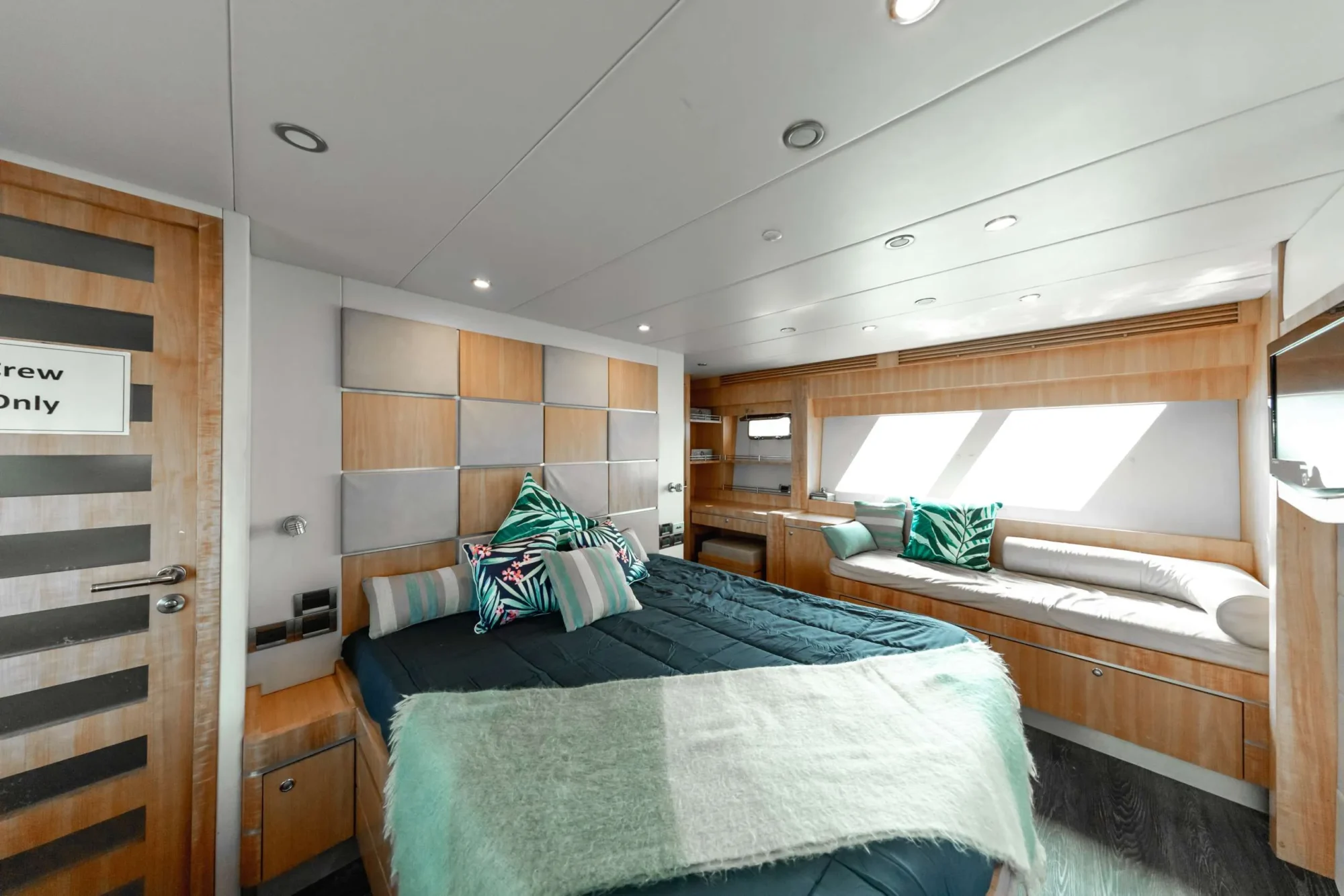 A modern bedroom with a double bed featuring green and black bedding, green striped and geometric-patterned pillows, and a green throw blanket. Wood paneling surrounds the room, and a striped wood door with a "Crew Only" sign is on the left. Experience ultimate comfort reminiscent of Luxury Yacht Rentals Sydney.