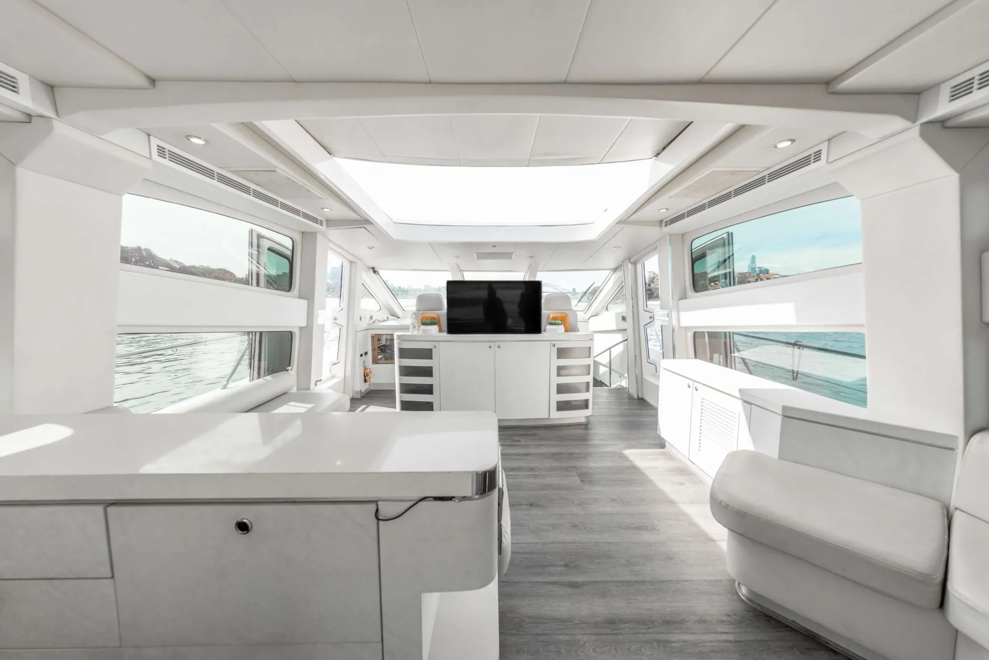 The image shows the interior of a modern, luxurious yacht. The space features sleek white furnishings, large windows providing ample natural light, a flat-screen TV in the center, and a stylish, minimalist design with a view of the water outside—perfect for Boat Parties Sydney The Yacht Social Club.