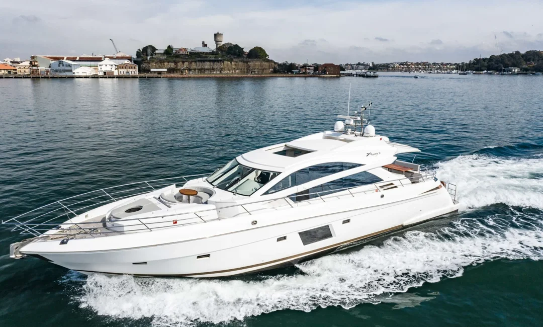 A white luxury yacht cruises through the calm waters with a coastal town and historic stone fortress visible in the background under a partly cloudy sky. The yacht, part of The Yacht Social Club Sydney Boat Hire, leaves a small trail of wake behind it as it moves forward.