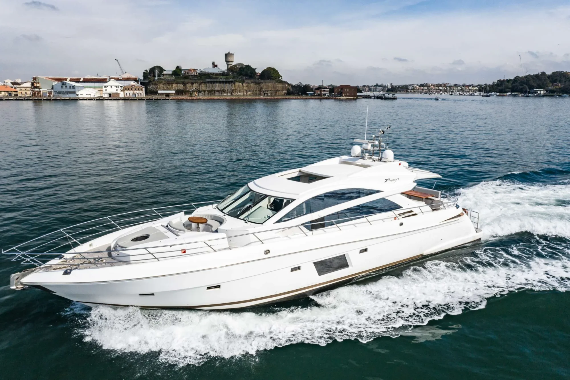 A white luxury yacht cruises through the calm waters with a coastal town and historic stone fortress visible in the background under a partly cloudy sky. The yacht, part of The Yacht Social Club Sydney Boat Hire, leaves a small trail of wake behind it as it moves forward.