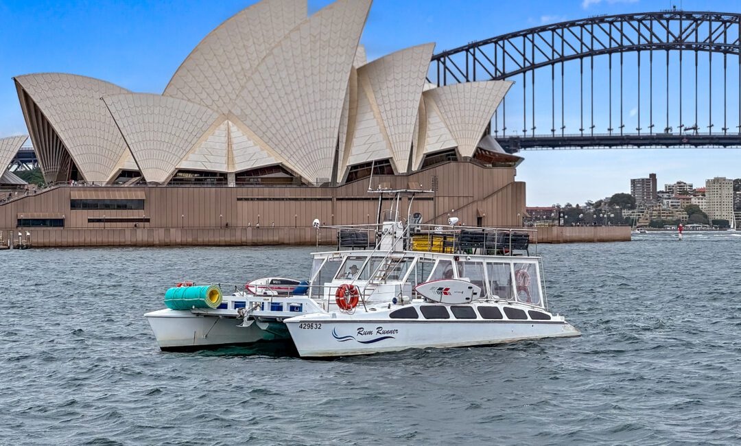 Rum Runner floats on the water in front of the iconic Sydney Opera House and Sydney Harbour Bridge. The sky is clear with a few clouds, and the urban skyline is visible in the background. The boat has several colourful objects on its deck.