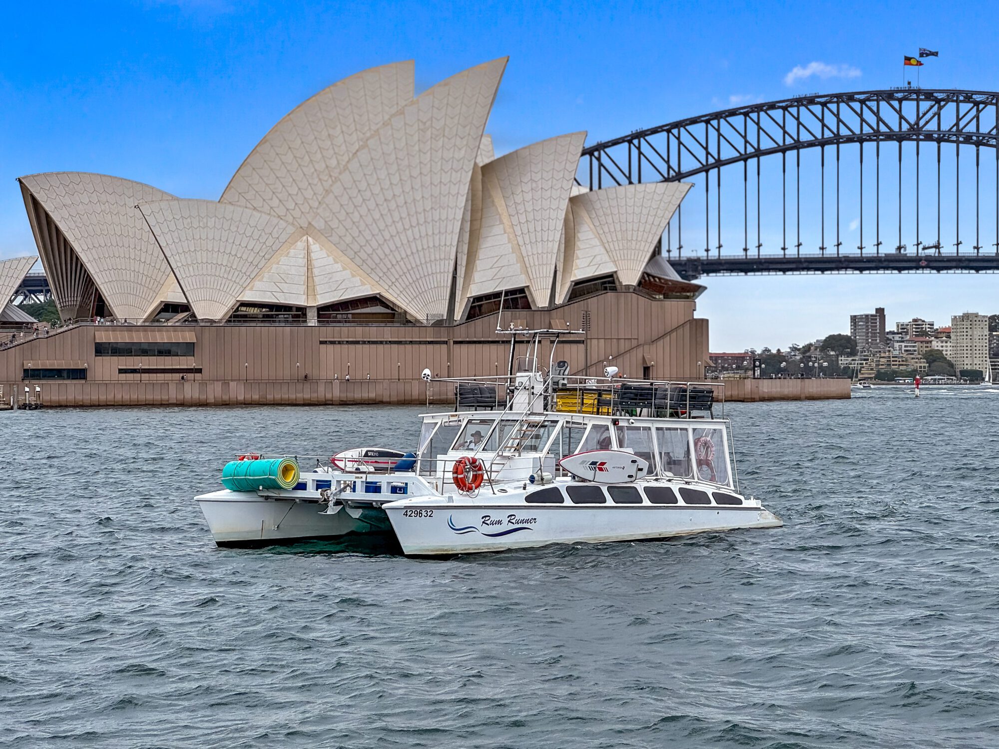 Rum Runner floats on the water in front of the iconic Sydney Opera House and Sydney Harbour Bridge. The sky is clear with a few clouds, and the urban skyline is visible in the background. The boat has several colourful objects on its deck.