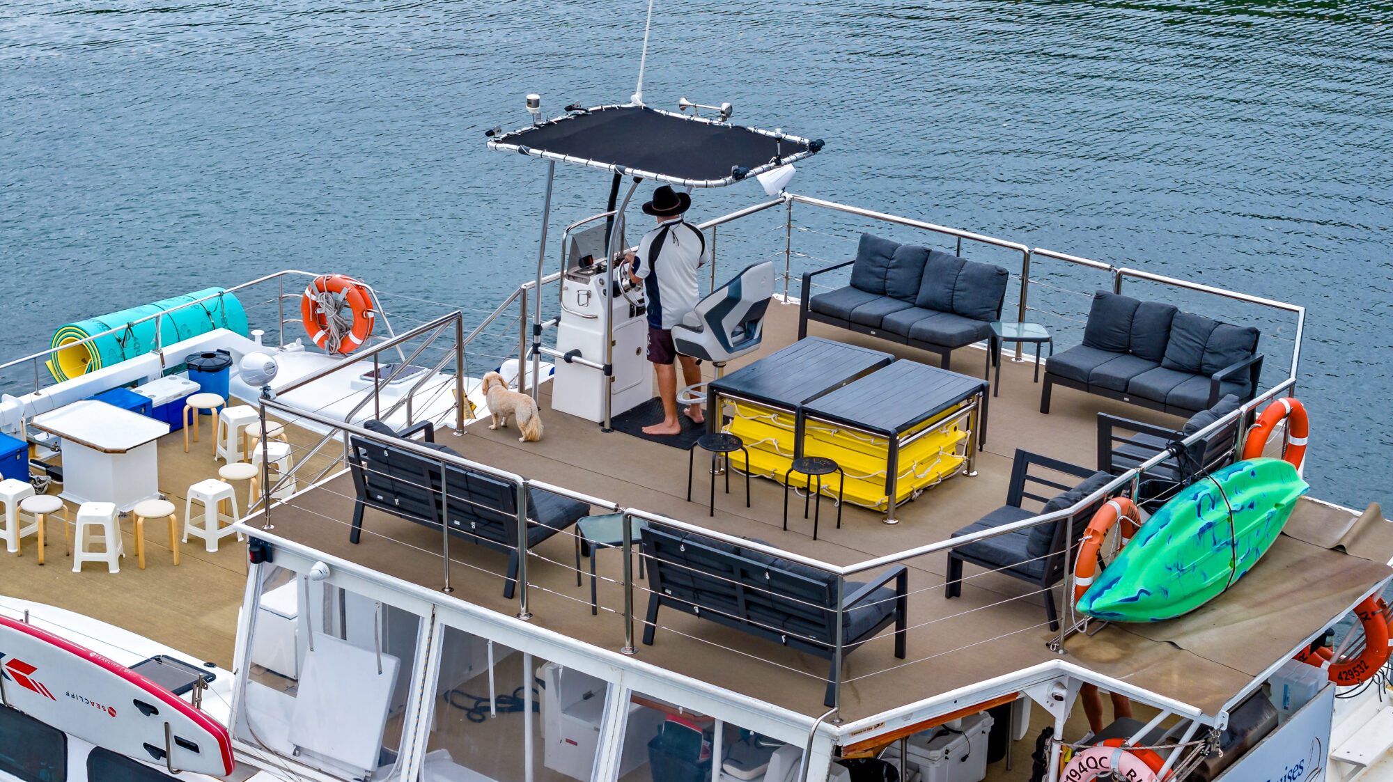 A man stands on the upper deck of a houseboat looking out at the water while holding a camera. He's accompanied by two small dogs. The deck is furnished with lounge chairs, a table, and kayaks. The water and shoreline set a serene backdrop.