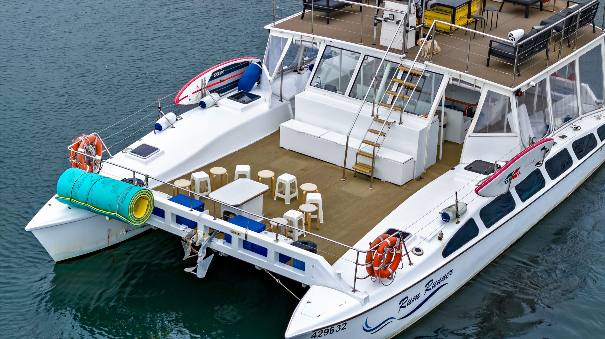 A white catamaran is docked on calm water. The boat features a spacious deck with several white chairs and tables, stairs leading to an upper level, and brightly colored life preservers and a rolled-up inflatable on the side. The boat's name is visible on the hull.