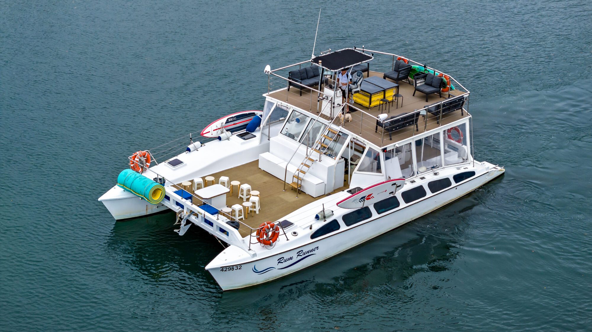 An overhead view of a large white catamaran on a body of water. The boat features multiple areas for seating, both open-air and partially covered, with lounge chairs and tables. Life preservers and surfboards are visible onboard.