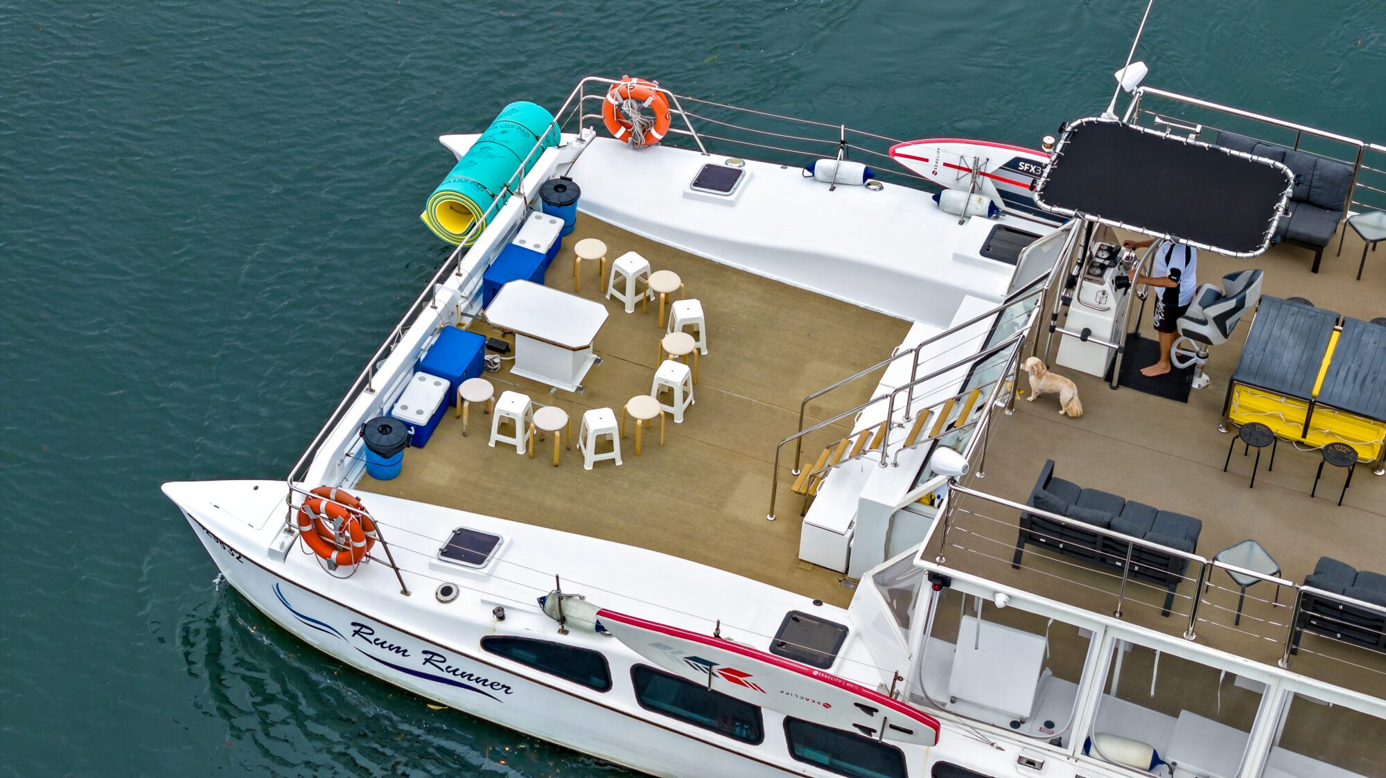 Aerial view of a boat named "River Runner" with a seating area on its deck. The deck has white tables and chairs, yellow towel racks, surfboards, and life rings. The boat is on calm water, and the image also shows parts of adjacent boats.