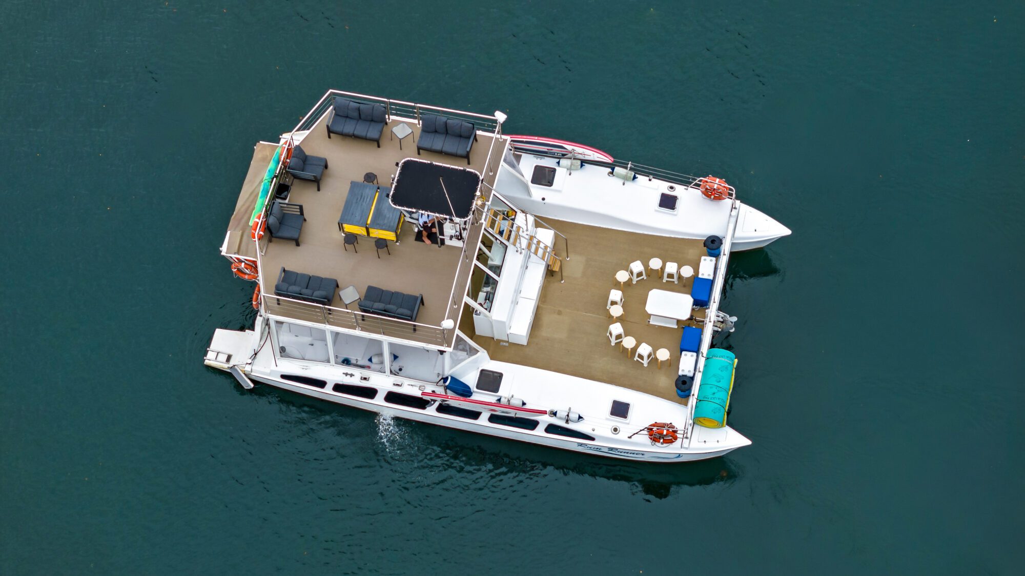 Aerial view of a multi-level catamaran boat sailing on clear blue water. The boat has outdoor seating areas on both levels, with tables and chairs arranged in circles on the deck. Several life preservers and umbrellas are also visible.