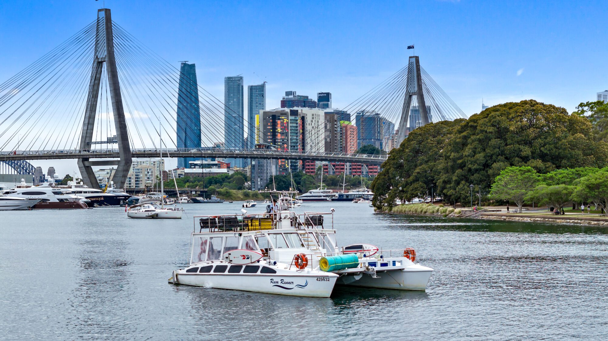 A scenic view of a waterfront featuring several boats docked in the water, with a prominent suspension bridge in the background. Modern skyscrapers and lush green trees provide a contrasting backdrop under a clear, blue sky.