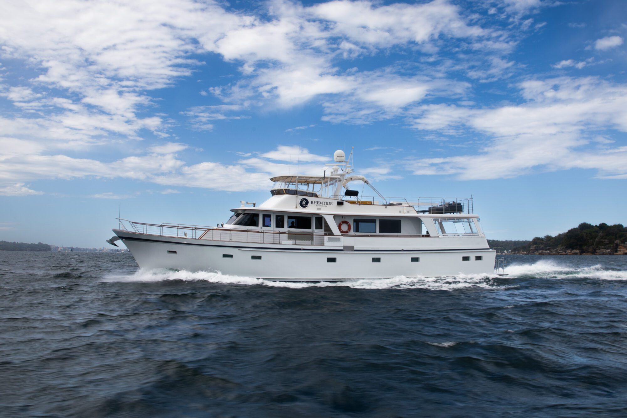 A white yacht moves swiftly through the ocean, creating waves in its wake under a bright blue sky with scattered clouds. The shoreline with trees is visible in the distance. The yacht appears modern and sturdy.