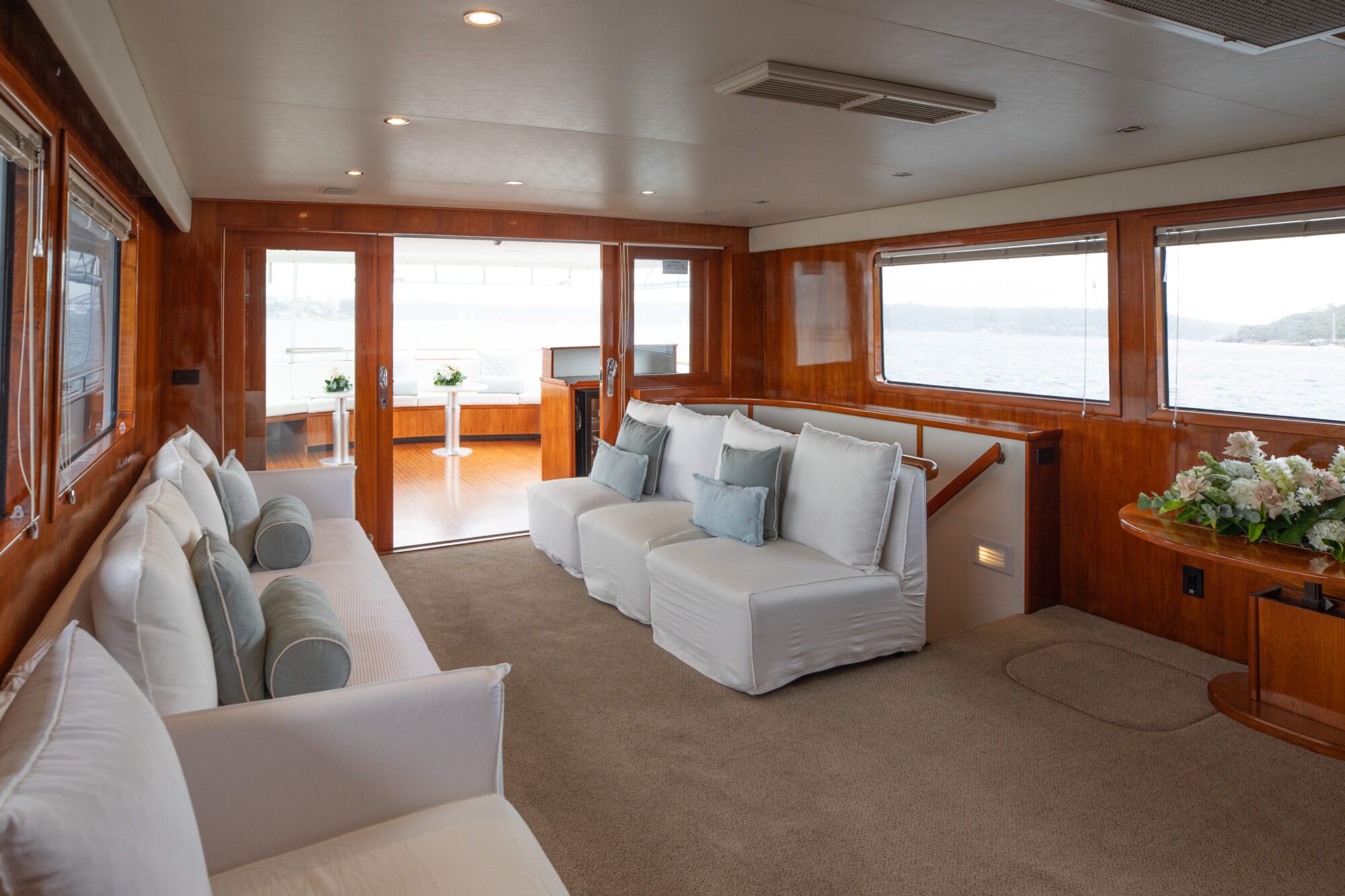 A luxurious yacht interior featuring a spacious living area with large windows offering scenic ocean views. The room has white upholstered sofas with light blue accent pillows, wooden paneling, and a vase of flowers on a small side table.