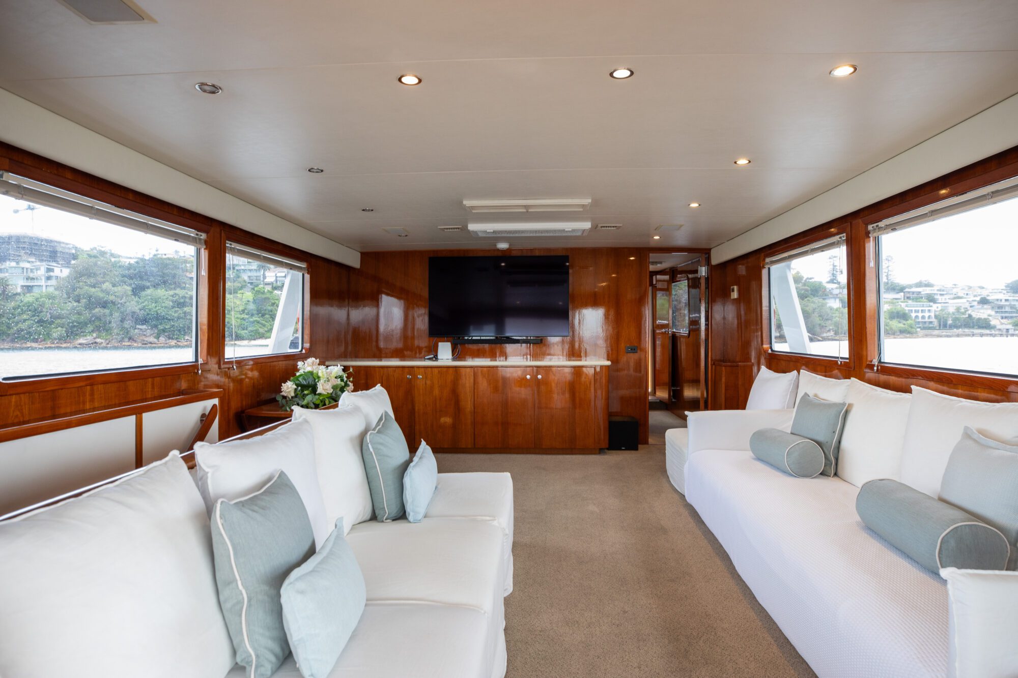 A spacious yacht lounge with large windows on either side, allowing ample natural light. The room features plush white sofas with blue throw pillows, a flat-screen TV mounted on a wooden wall panel, and a vase of flowers on a table in the corner.