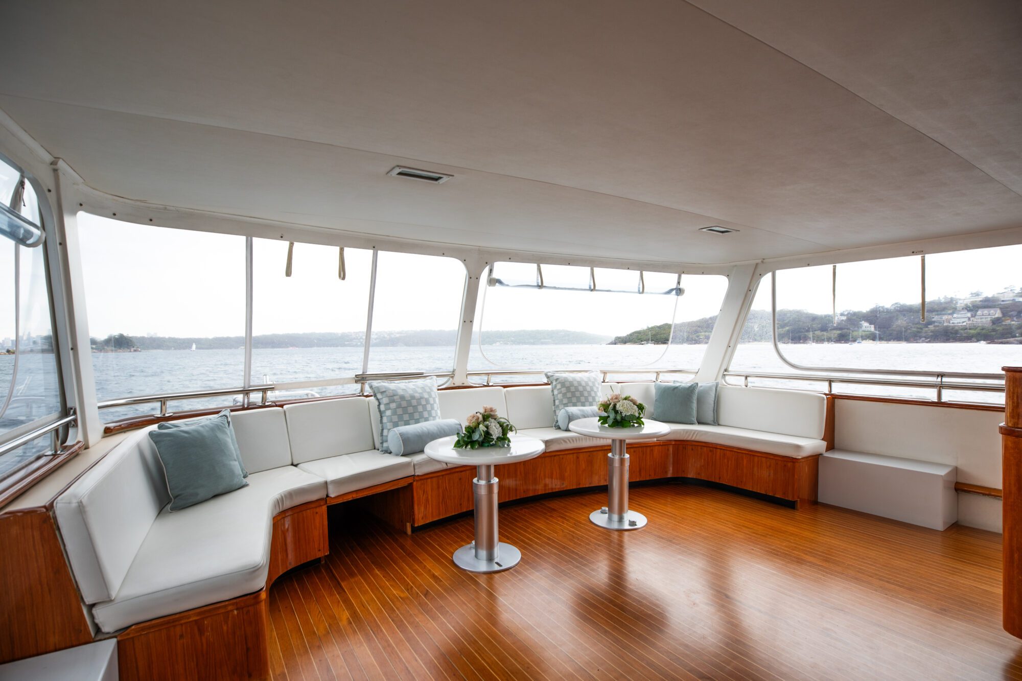 A spacious boat lounge with large windows offers a panoramic view of a calm body of water and distant shoreline. The interior features white cushioned seating arranged in an L-shape, wooden floor, and two small round tables adorned with flower arrangements.