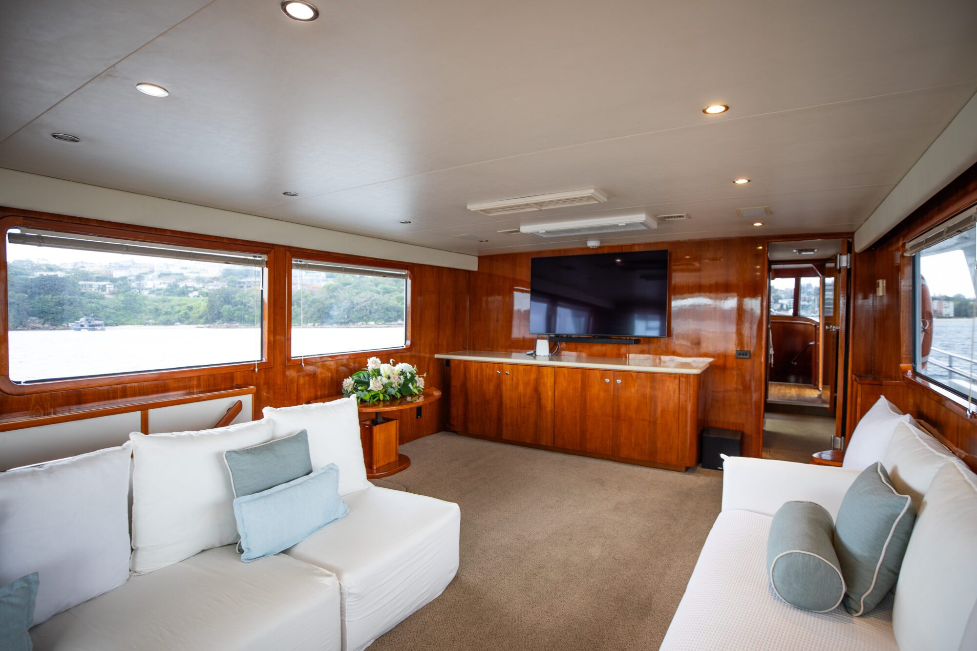 Luxurious yacht interior with plush white sofas and light blue cushions. The space features large windows, wooden paneling, recessed lighting, a large flat-screen TV, and a cabinetry unit. Scenic views of water and greenery are visible through the windows.