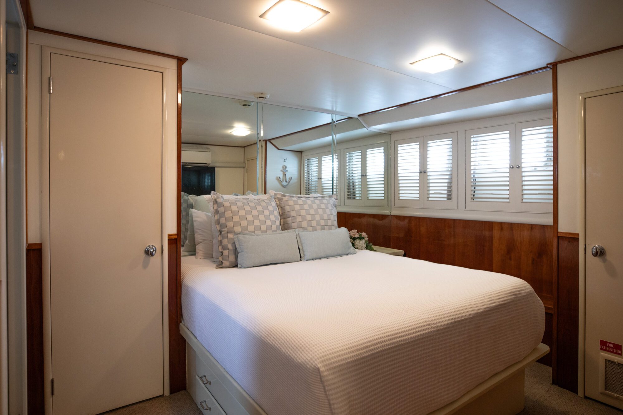 A cozy bedroom features a neatly made bed with white linens and multiple pillows. The room has wood-paneled walls with mirrors, a closet, and shuttered windows. Soft lighting adds a warm ambiance, and a small vase of flowers decorates the bedside.