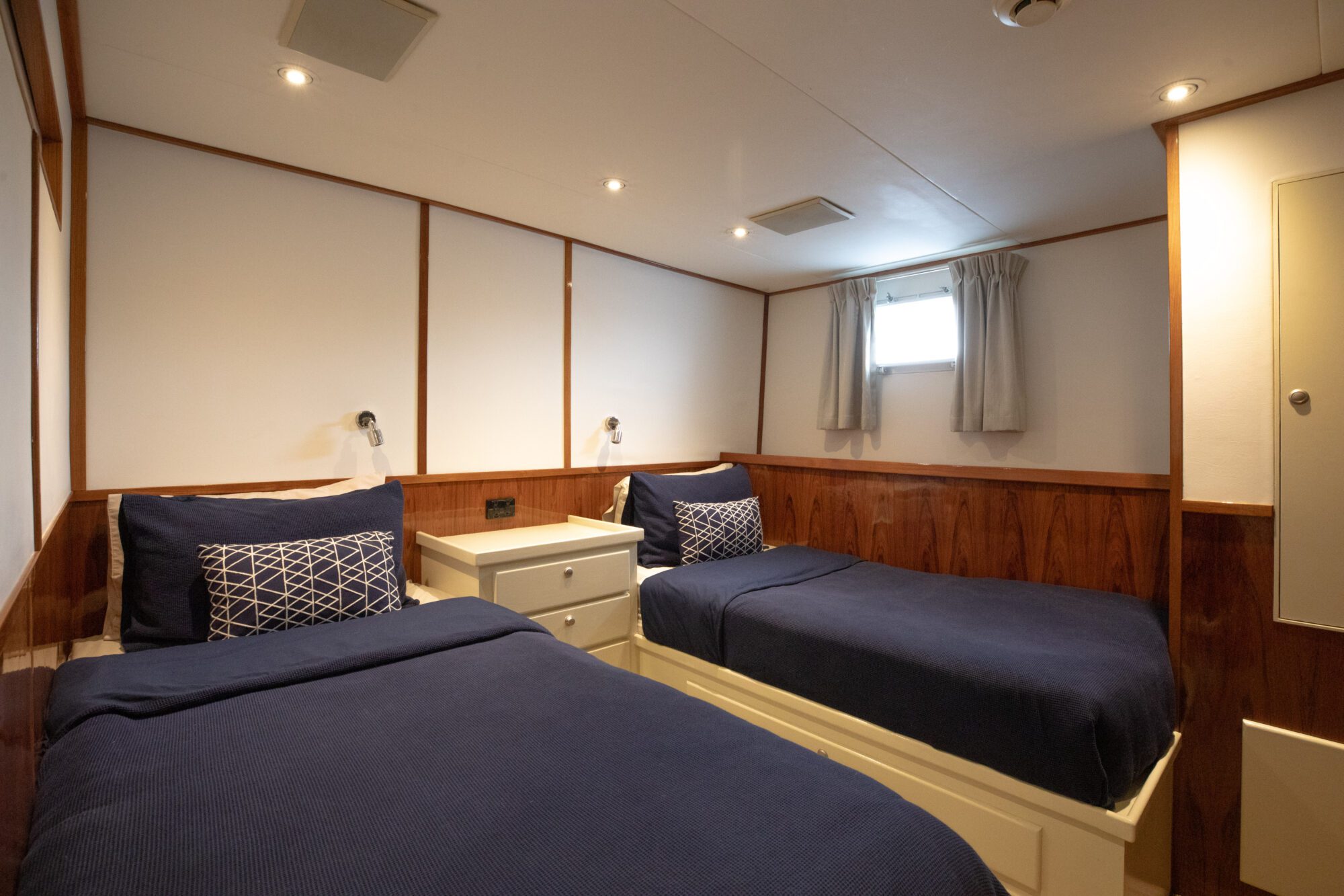 A cozy room features two twin beds with dark blue bedding, separated by a small white nightstand. The walls are wood-paneled with white accents, and a small window with closed curtains lets in light. Recessed lighting provides additional illumination.