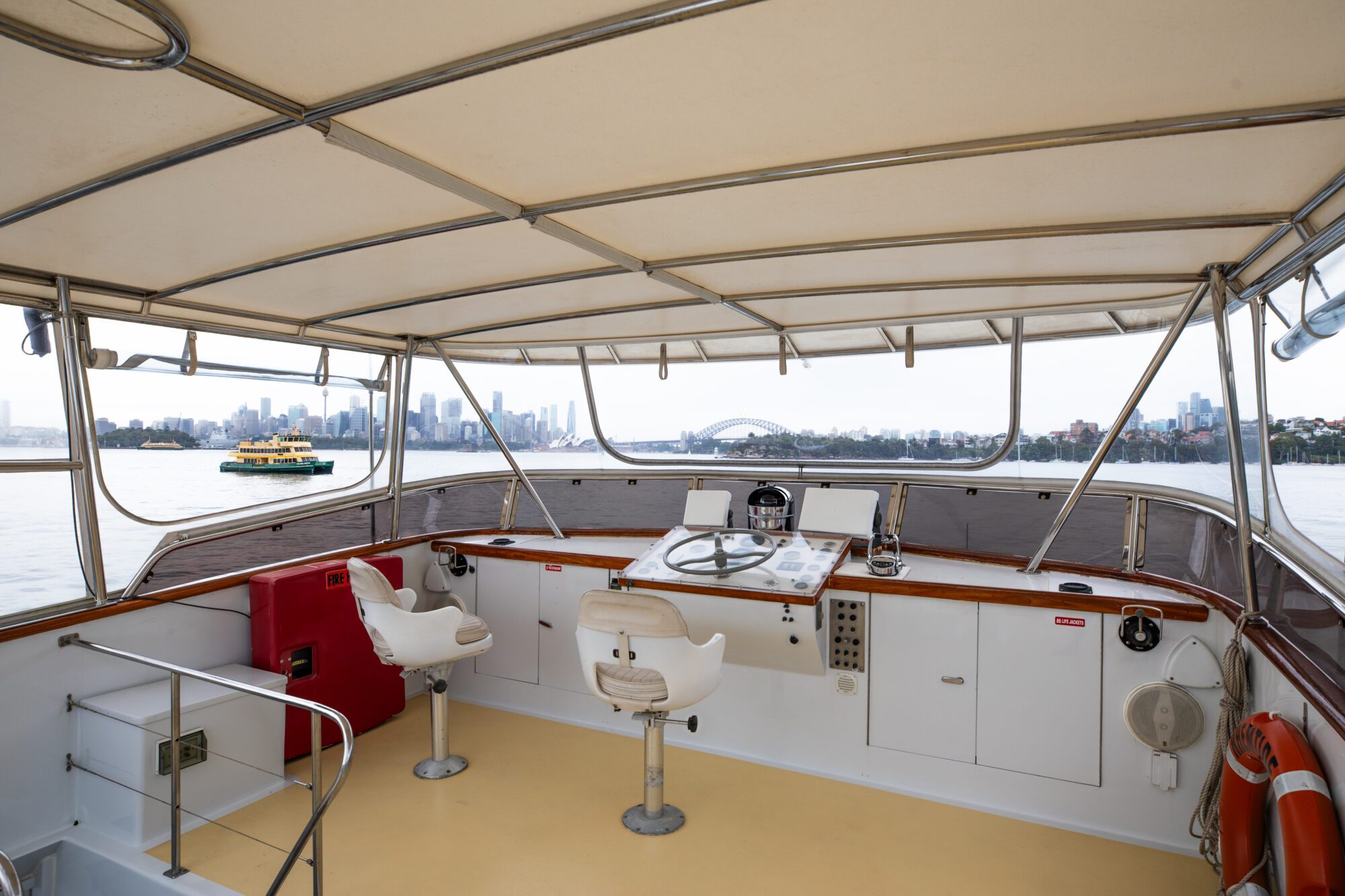 The image shows the upper deck of a boat with two white swivel chairs facing the helm. The boat is covered with a canopy, and there is a view of a harbor with city buildings and a bridge in the background. A green and yellow ferry is visible on the water.