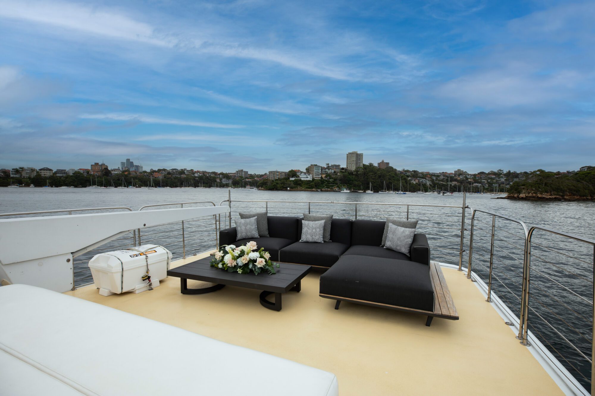 The deck of a boat with a comfortable seating area featuring a black sofa and a coffee table with a flower arrangement. The boat is docked near a cityscape with buildings and trees visible in the background under a partly cloudy sky.