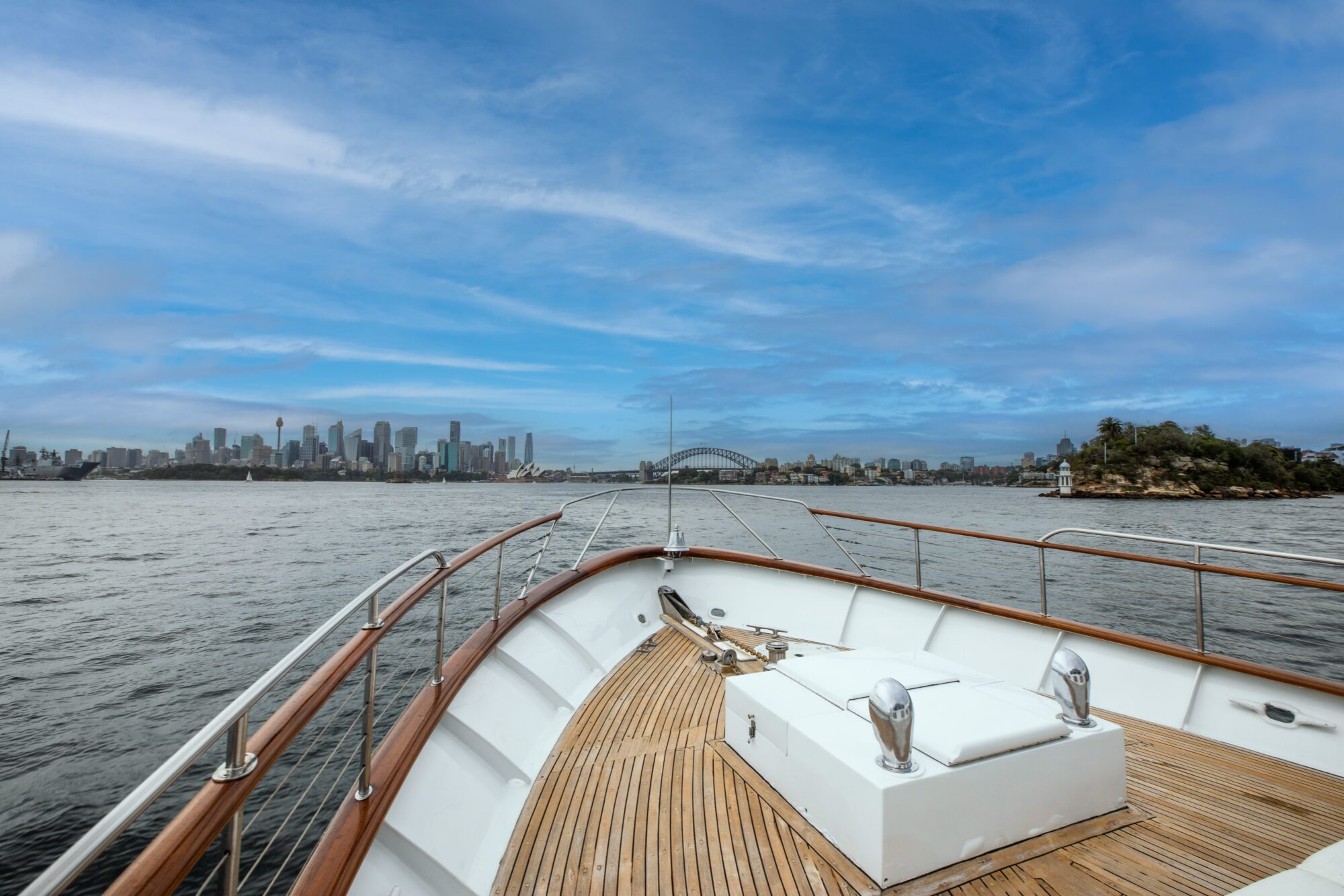 A view from the bow of a boat looking toward a city skyline in the distance, with calm water surrounding and a partly cloudy sky above. The deck has wooden planks and stainless steel fixtures, and the cityscape includes a prominent bridge.