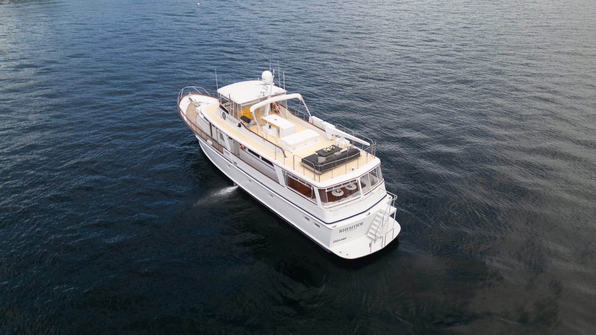 A white yacht cruises on a calm, expansive body of water. The vessel has multiple decks, a seating area at the rear, and a radar on top. The water is dark and reflective, stretching into the distance, with no land visible nearby.