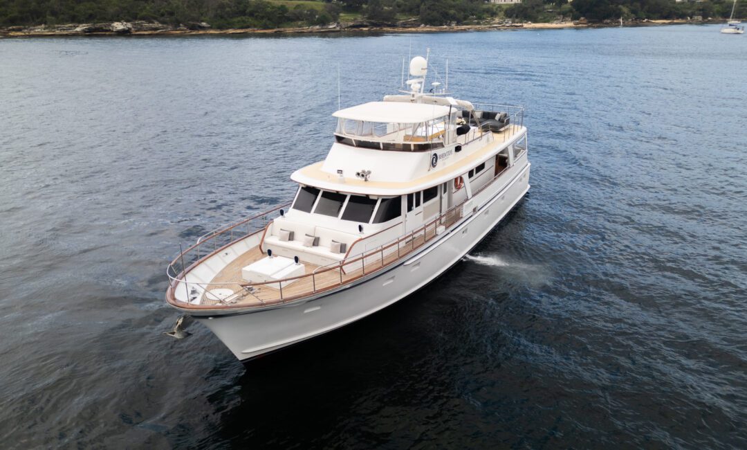 Aerial view of a large white motor yacht cruising on a calm body of water near a forested shoreline. The yacht has multiple decks, antennas, and a beige roof. The water is dark blue, and the shoreline in the background features dense trees and some rocky areas.