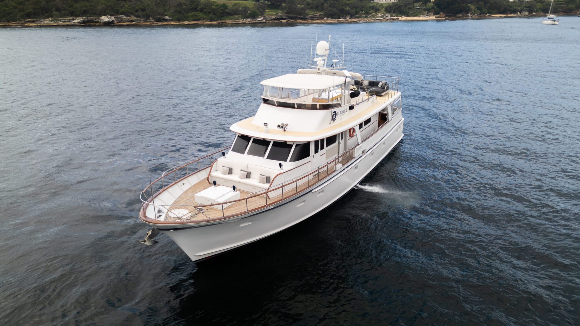 Aerial view of a large white motor yacht cruising on a calm body of water near a forested shoreline. The yacht has multiple decks, antennas, and a beige roof. The water is dark blue, and the shoreline in the background features dense trees and some rocky areas.