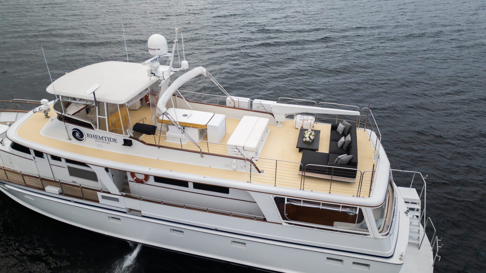 Aerial view of a luxury yacht named "TIME TIDE" sailing on calm waters. The upper deck features a shaded area with seating, a dining table with flowers, and a sunbathing area. The yacht has a sleek white design, and the surrounding water appears serene and vast.