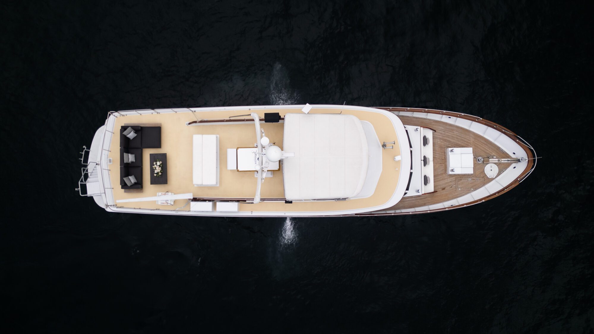 Aerial view of a yacht on dark water. The yacht features a spacious deck with seating areas, sunbeds, and a control station toward the front. The vessel is moving, evident from the white wake behind it.