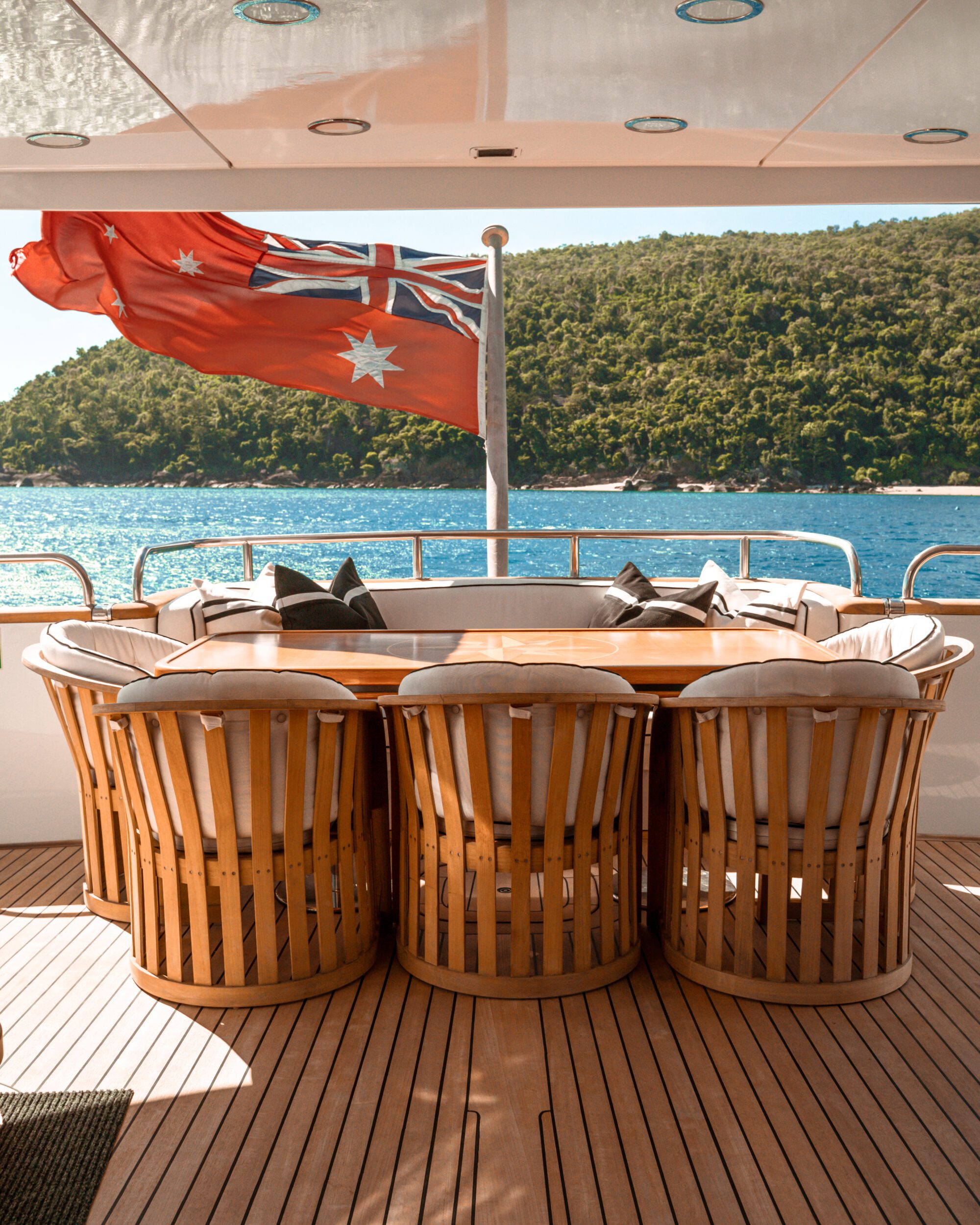 A wooden deck on a yacht features a circular table surrounded by cushioned chairs. An Australian flag waves at the back, with an island covered in greenery visible across the sparkling blue sea.