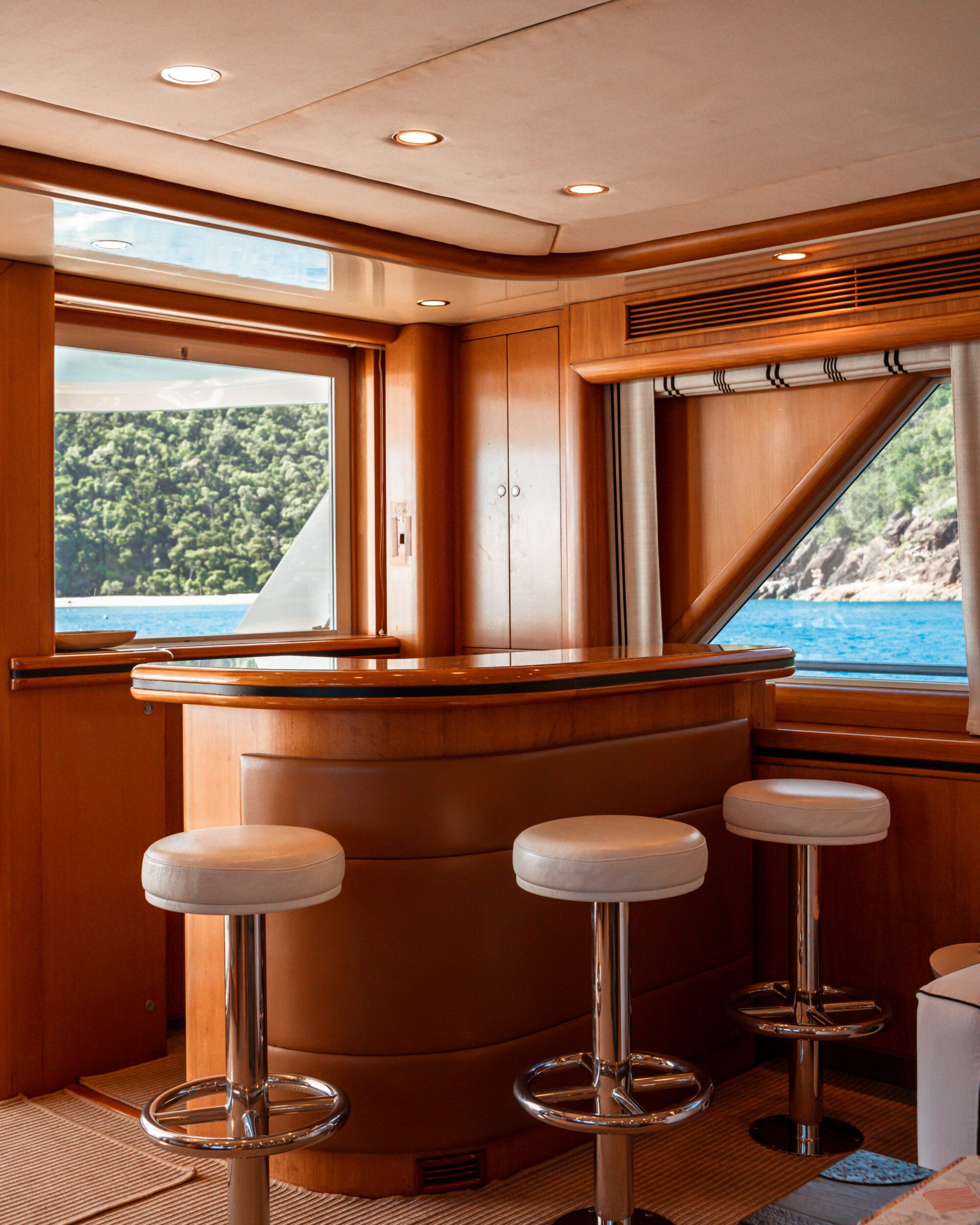 Luxurious yacht interior with a polished wooden bar and three white cushioned stools. Large windows reveal a scenic ocean view and lush green hills in the background. Warm lighting creates an inviting atmosphere.