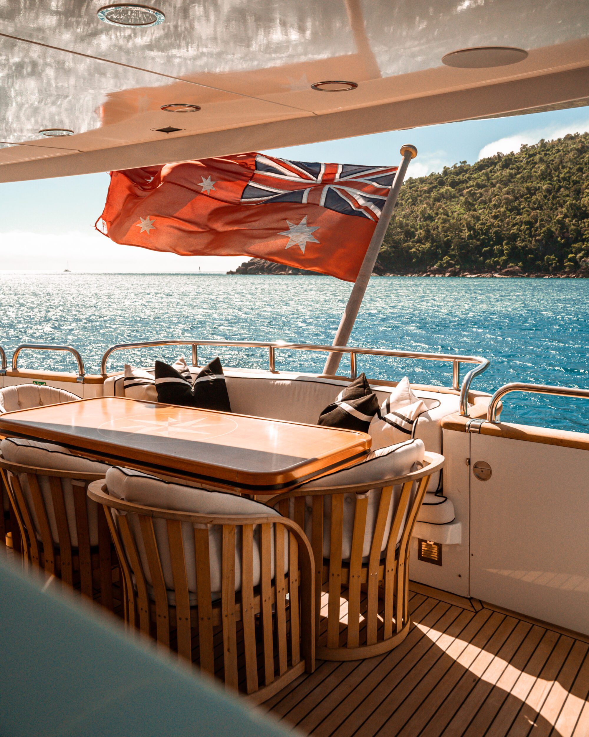 A luxurious yacht deck features a polished wooden dining table surrounded by chairs. An Australian flag waves in the breeze, with a serene blue ocean and a green, tree-covered island visible in the background.