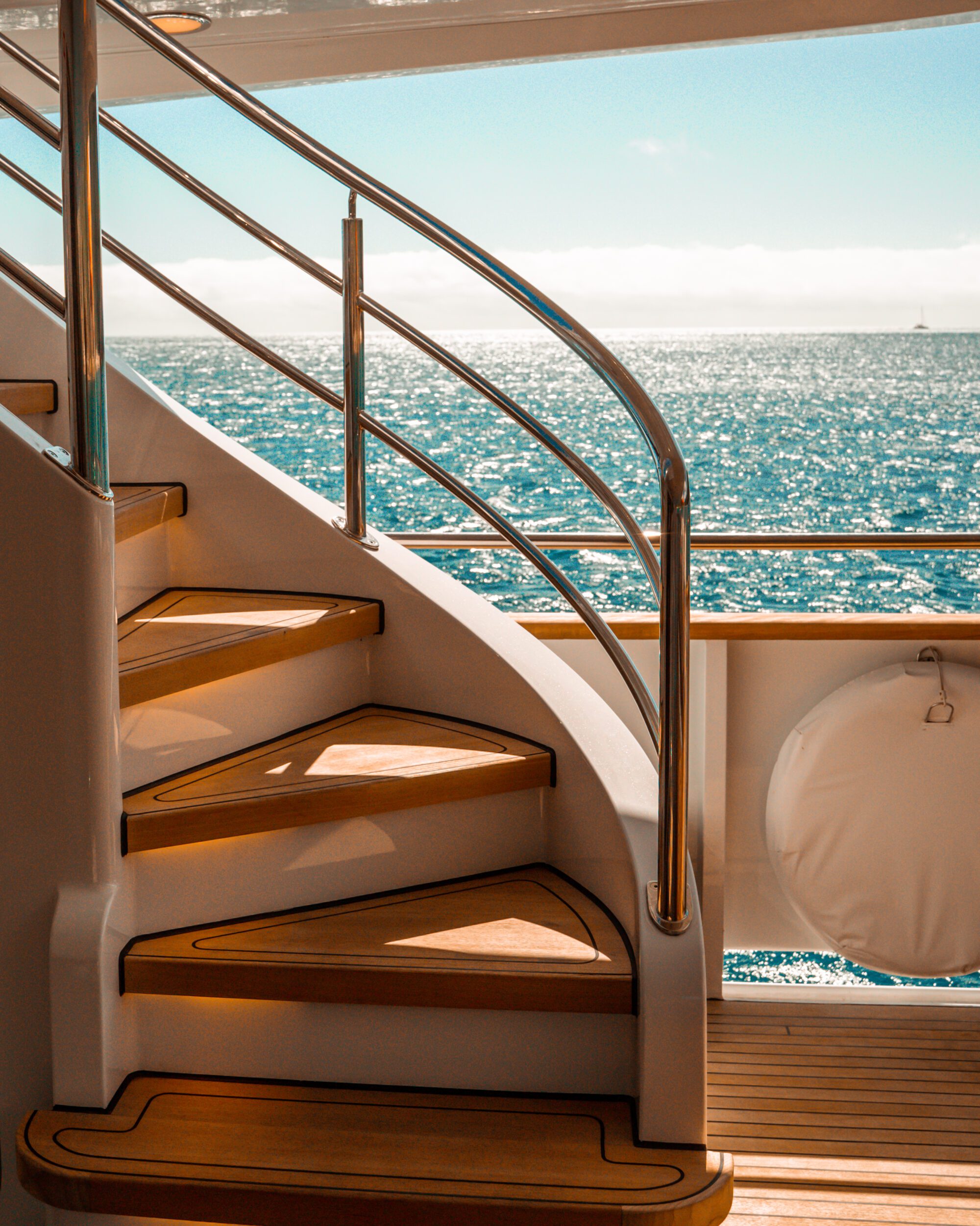 Wooden stairs with metal railings on a boat lead upward, overlooking a calm, turquoise ocean under a clear blue sky. A life preserver is attached to the railing.