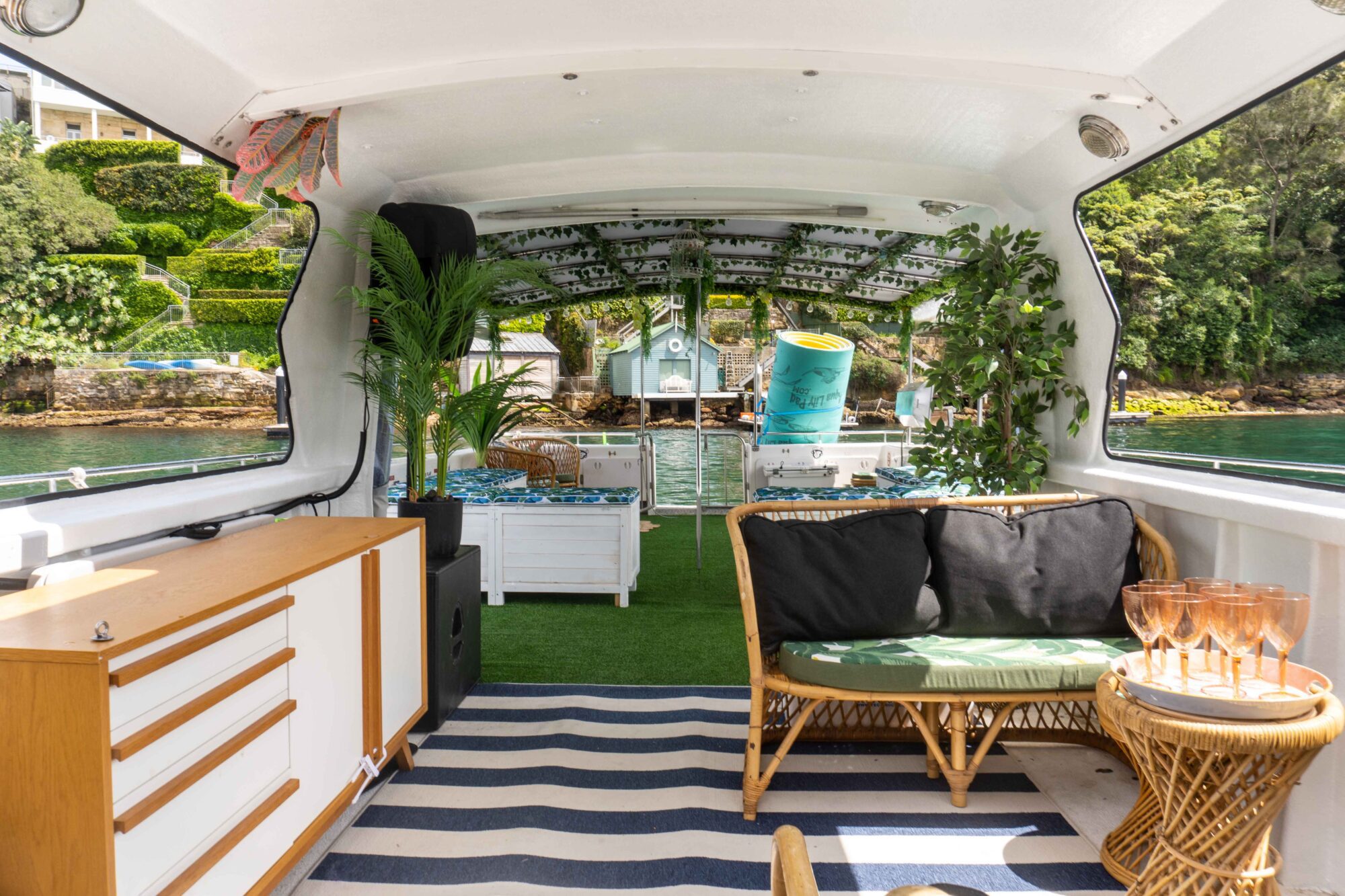 Interior of a boat adorned with wicker furniture, a striped rug, and lush green plants. Sunlight pours in through large windows, revealing a scenic waterfront view with greenery outside. A small cabinet and dining table with glasses complete the cozy setup for The Yacht Social Club Event Boat Charters.