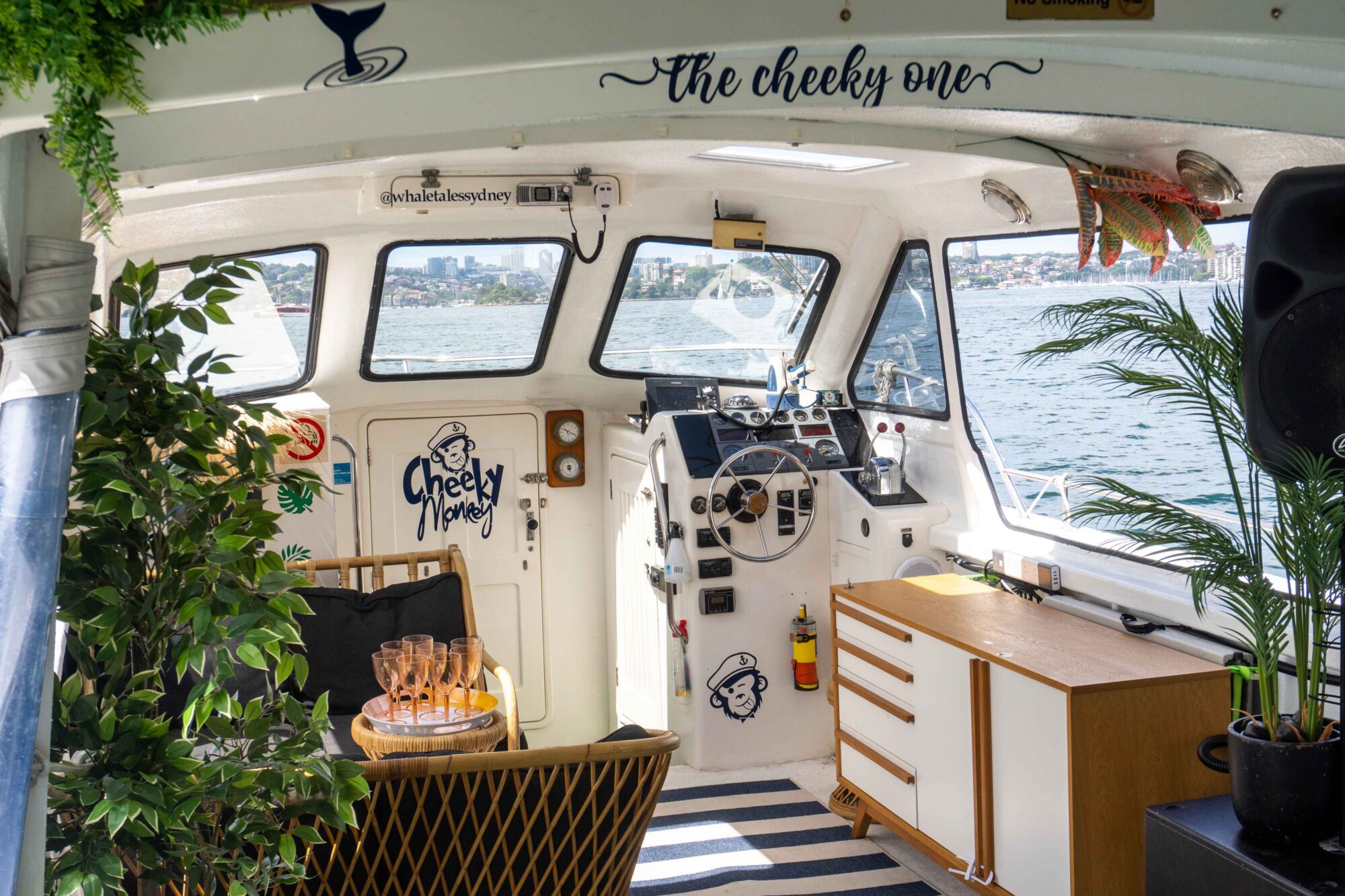 Interior of a cozy boat named "The Cheeky One," decorated with plants, a striped rug, and a wicker chair. The helm features navigation instruments and drinks on a nearby table. Large windows offer a scenic view of the harbor and cityscape—perfect for The Yacht Social Club Event Boat Charters.