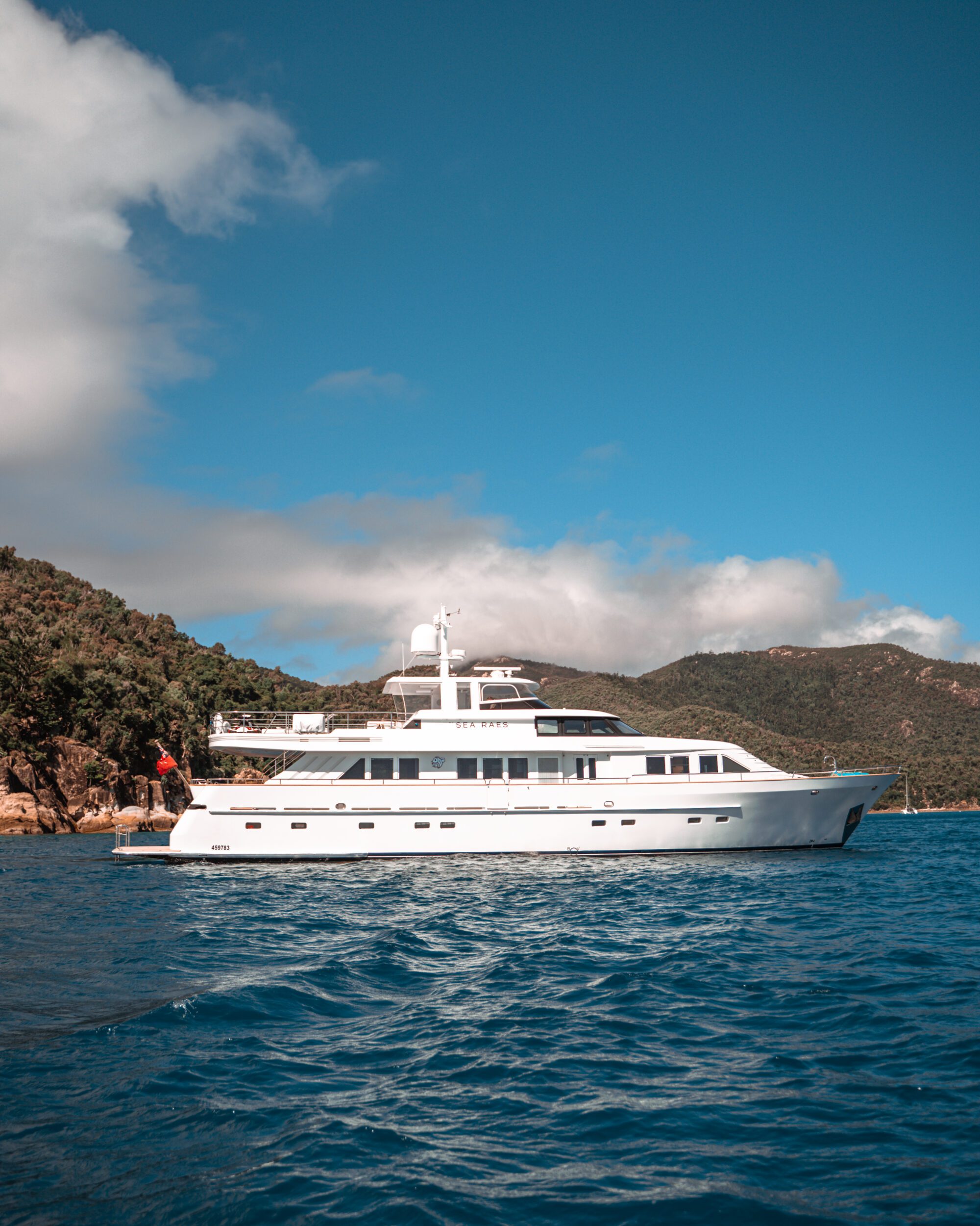 A large white yacht is anchored on a clear blue body of water, with lush green hills and a partly cloudy sky in the background. A person in red clothing stands on the yacht's deck.