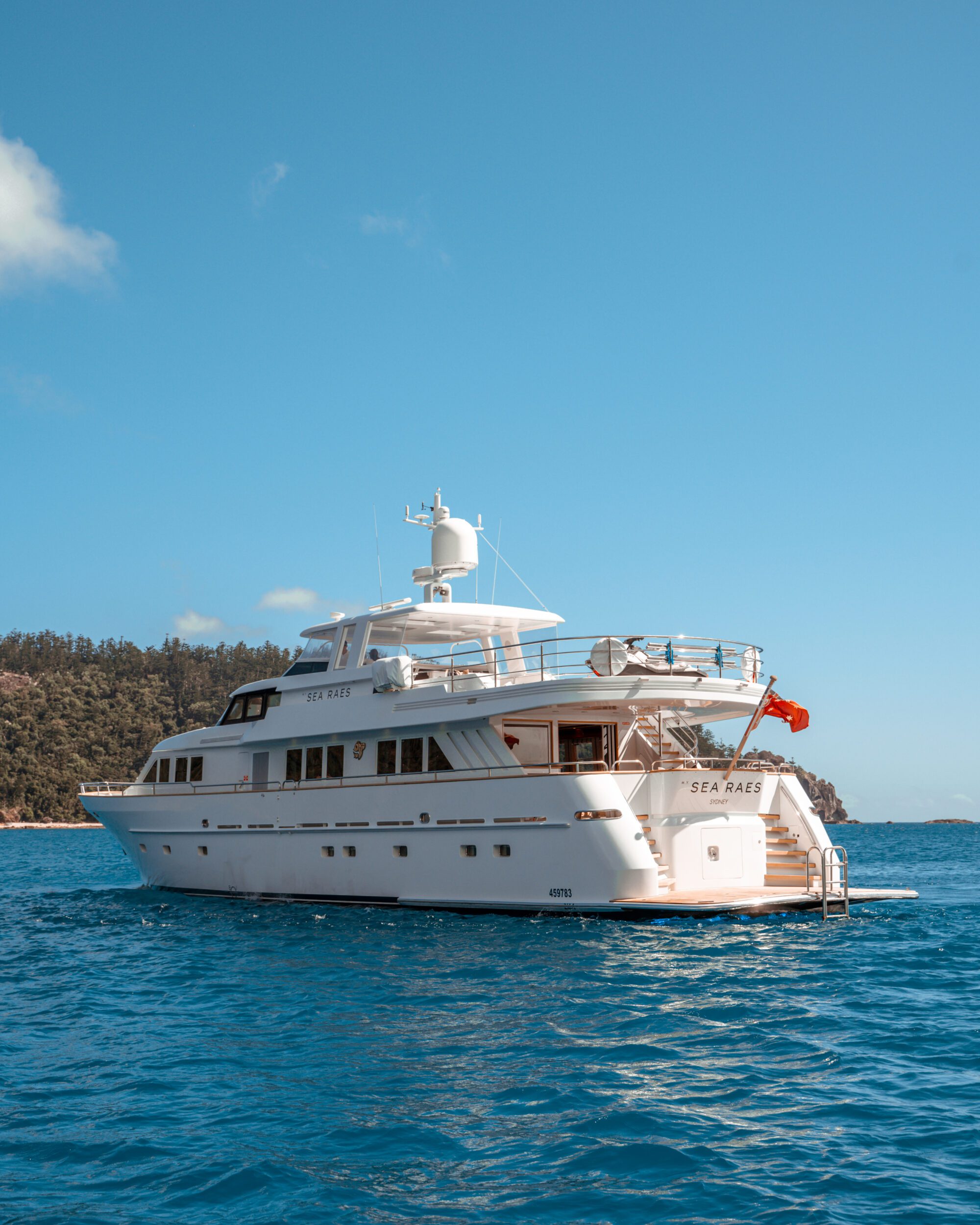 A luxurious white yacht named "Sea Raye" anchored on a clear blue ocean. The vessel has multiple decks and an open area at the stern. A forested shoreline is visible in the background under a bright blue sky.