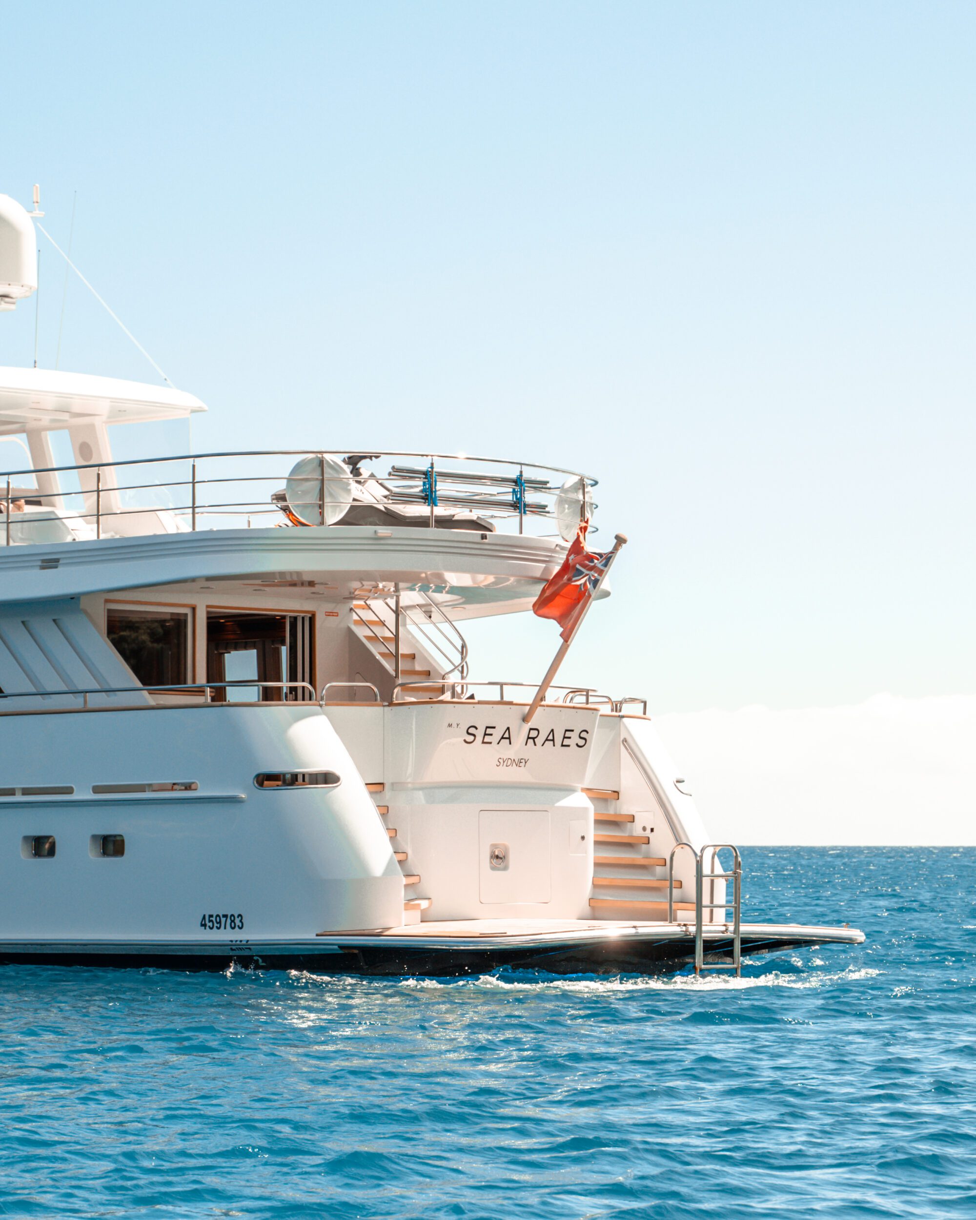 A luxury yacht named "Sea Raes" is anchored in calm, clear blue waters on a sunny day. The vessel features multiple decks and a small flag at the stern. The yacht's home port, "Sydney," is written below the name. Registration number 459783 is visible.