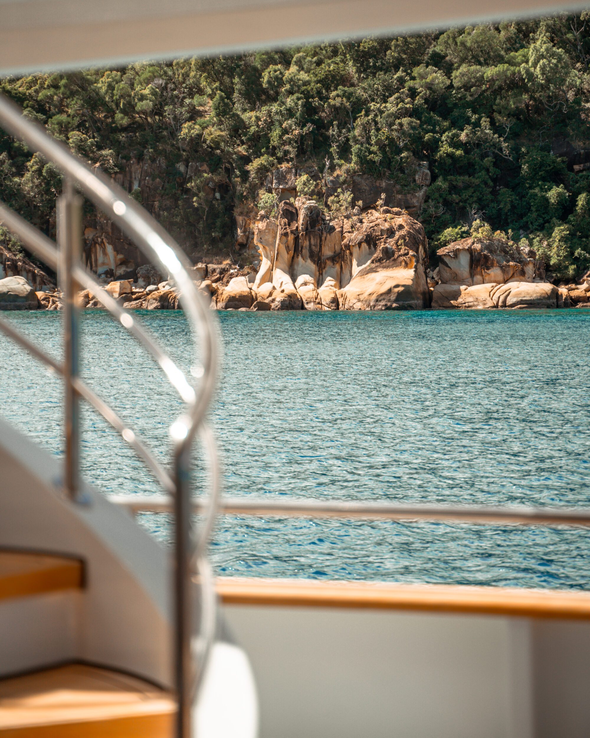 View from a boat, showing a staircase railing in the foreground and calm, turquoise water with a rocky, tree-lined shore in the background.