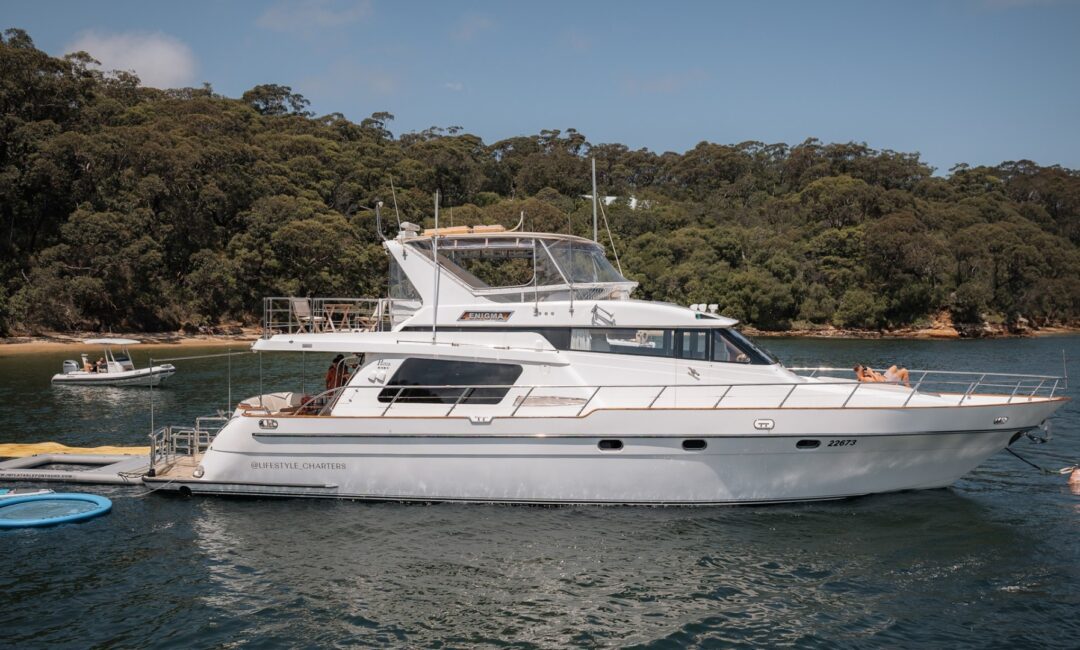 A white luxury yacht is anchored in a calm bay surrounded by dense, green forest. The boat has multiple decks, and people can be seen relaxing on board. A smaller boat is tethered nearby. The water is serene, reflecting the clear blue sky.