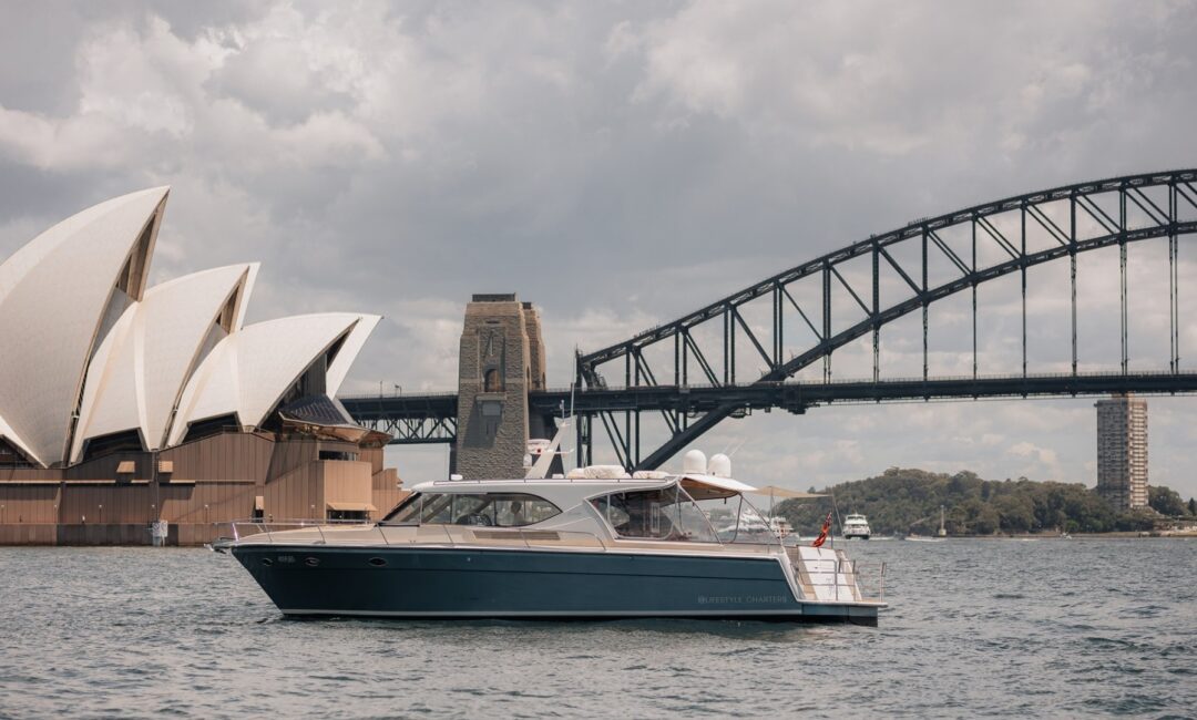 A luxury boat on Sydney Harbour is cruising near the iconic Sydney Opera House and the Sydney Harbour Bridge on an overcast day. The boat is sleek and modern, with a few people seen on board enjoying the view.