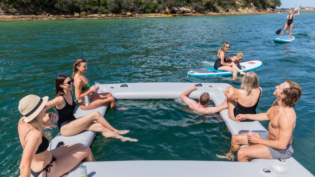 A group of people in swimsuits enjoy a sunny day on the water, lounging on and around a large inflatable platform with an open center. In the background, others paddleboard. The water is calm, and the shoreline is covered with trees and bushes.
